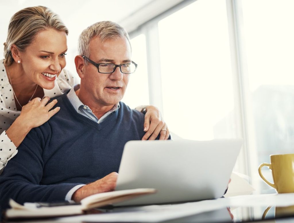 A happy, senior couple reviews information on a laptop together. The woman is looking over the man's shoulder, with her hand on his arm, and both are smiling.